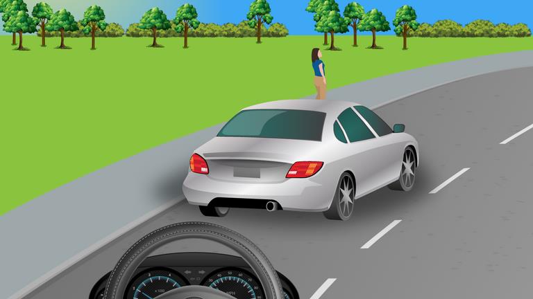 Driver's view following a silver car on a road; a person stands on the left verge farther ahead.
