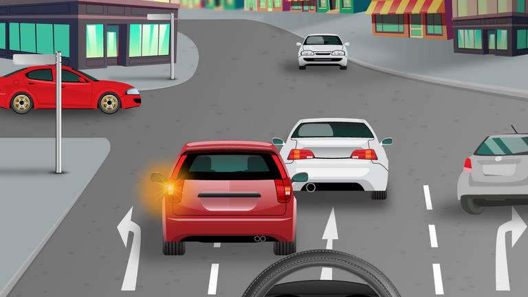 Driver's view approaching a junction behind a red car with an amber rear lamp and a white car; painted arrows mark the lanes.