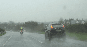 Photograph through a rain-covered windscreen, with a dark car ahead and a motorcycle farther to the left on a wet road.