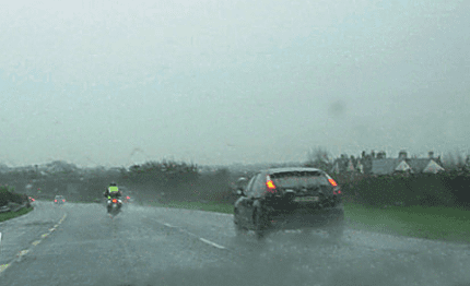 Photograph through a rain-covered windscreen, with a dark car ahead and a motorcycle farther to the left on a wet road.