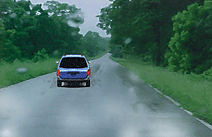 Photograph of a blue car ahead on a wet tree-lined road, with water on the road surface.
